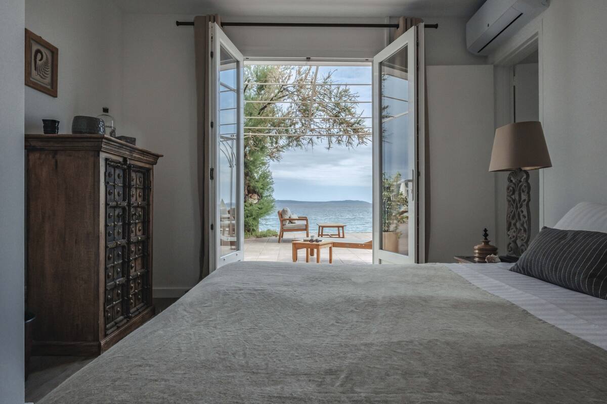 A bedroom with a large bed, a wooden cabinet, a nightstand, a lamp, and a view of the sea through an open door.