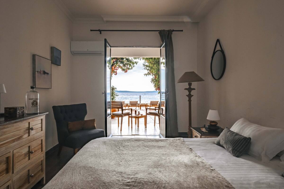 A bedroom with a bed, a chair, a dresser, a lamp, and a view of the sea through an open door.