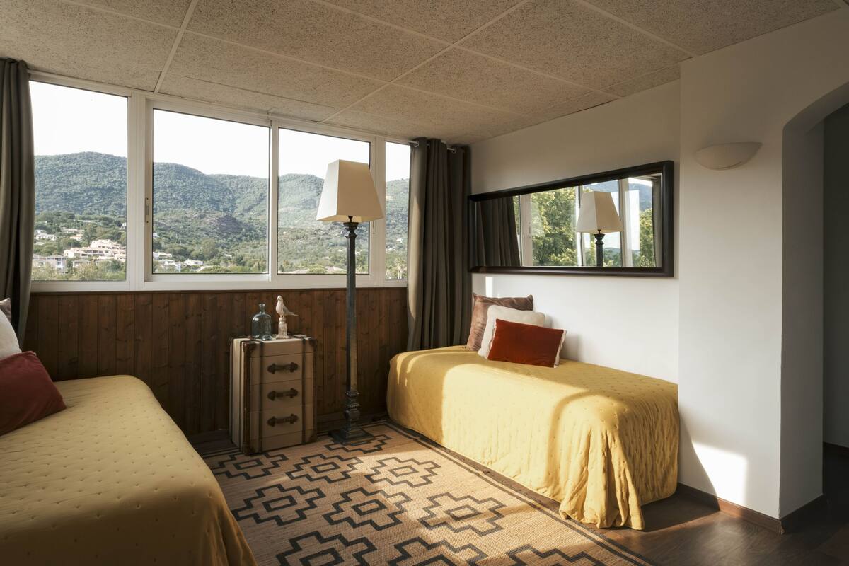 A hotel room with two beds, a window with a view of hills, a lamp, and a dresser.