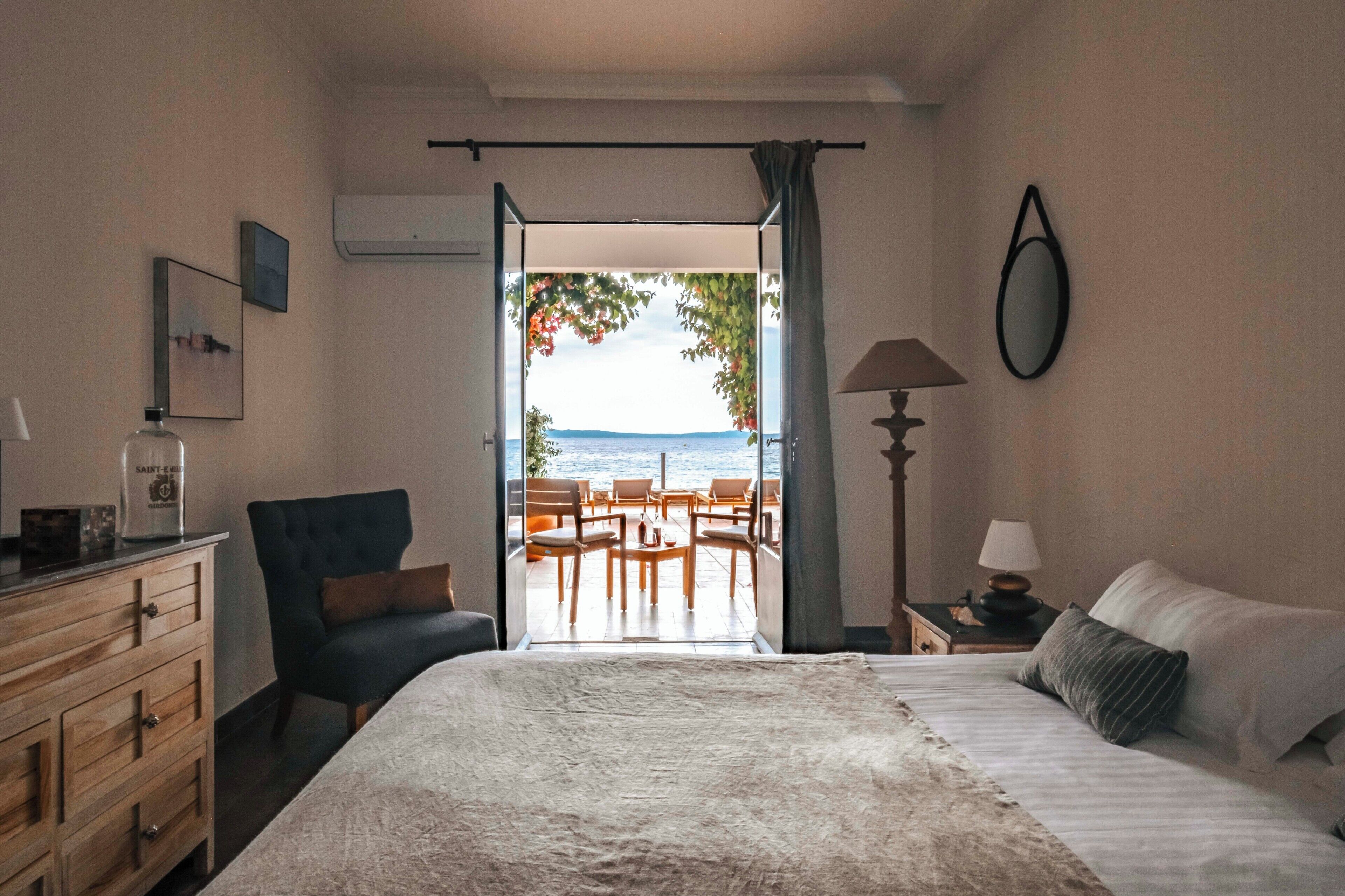 Un dormitorio con cama, una silla, tocador, lámpara y vista al mar a través de una puerta abierta.