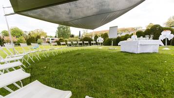 Un espacio al aire libre con césped, sillas blancas plegables dispuestas en filas, una mesa cubierta con mantel blanco y una pantalla o tablero blanco.