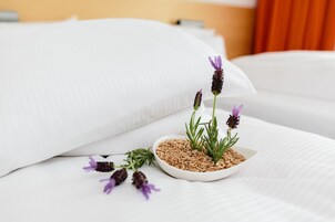 Una cama bien hecha con sábanas blancas, un pequeño cuenco blanco con semillas y ramitas verdes, y flores moradas sobre la cama.