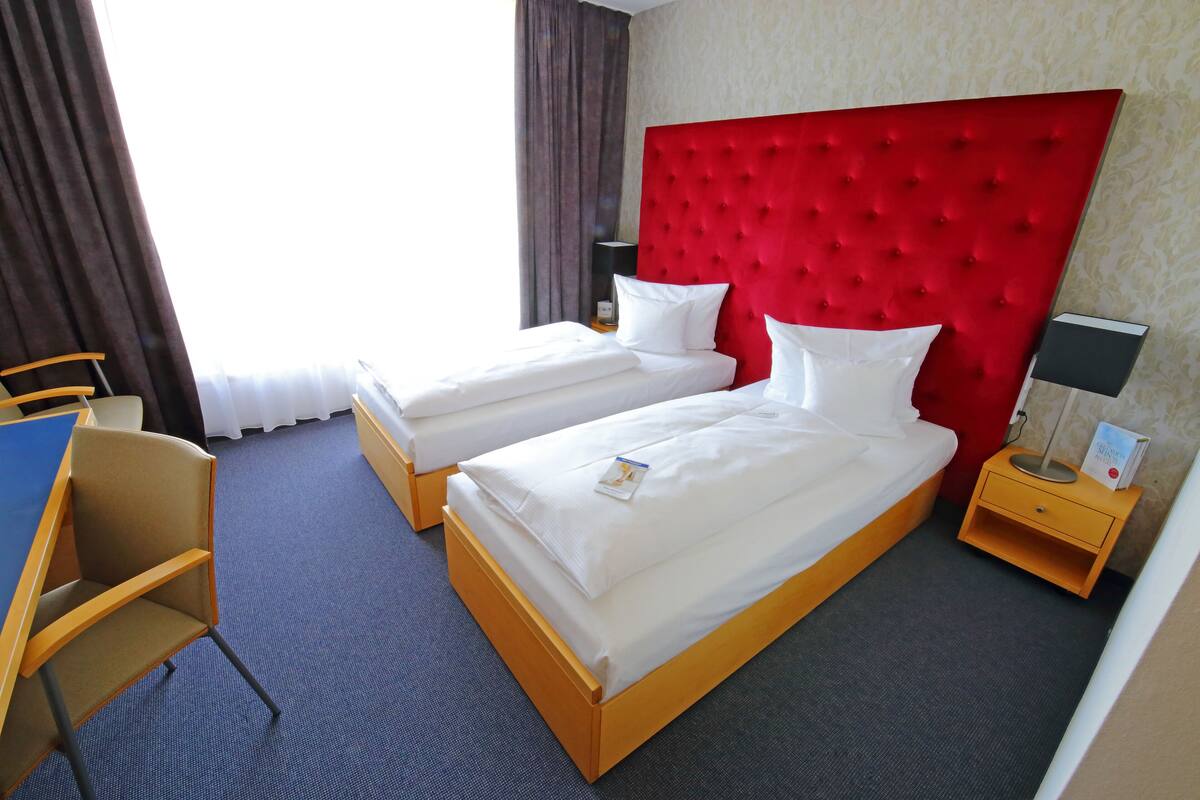 A hotel room with two single beds, a desk, a chair, a nightstand with a lamp, and a window with curtains.
