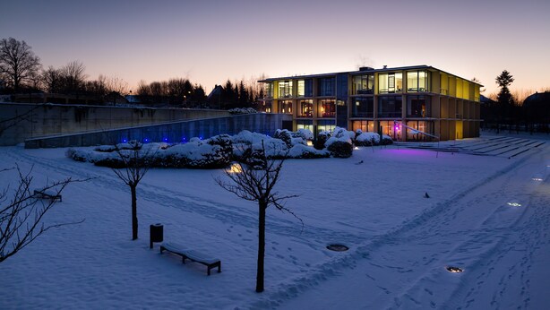 A modern building with a glass facade, surrounded by snow-covered ground and a few trees.