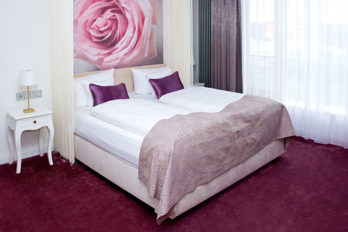 A hotel room with a large bed, a nightstand, a wall-mounted lamp, and a window with sheer curtains.