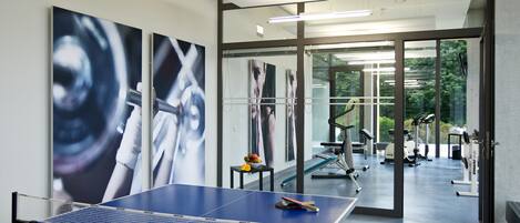 Mesa de ping pong en un espacio interior moderno con ventanas amplias, equipo de ejercicio y obras de arte en las paredes.