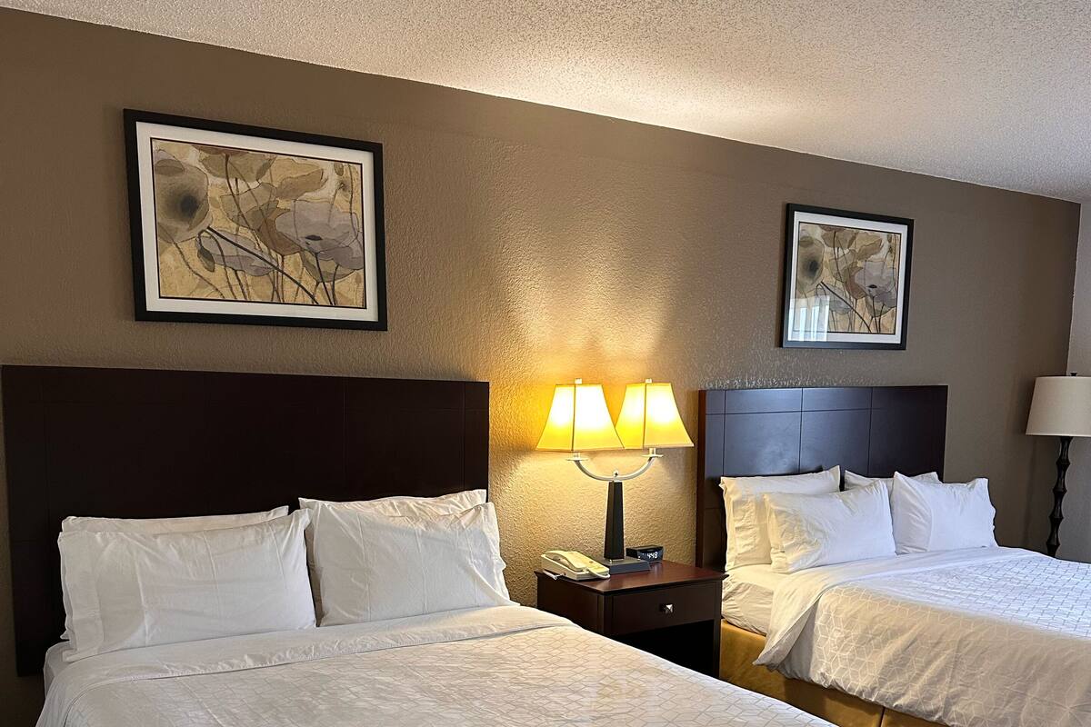 A hotel room with two beds, each with white linens and a headboard. There are two wall-mounted lamps and two framed floral artworks.