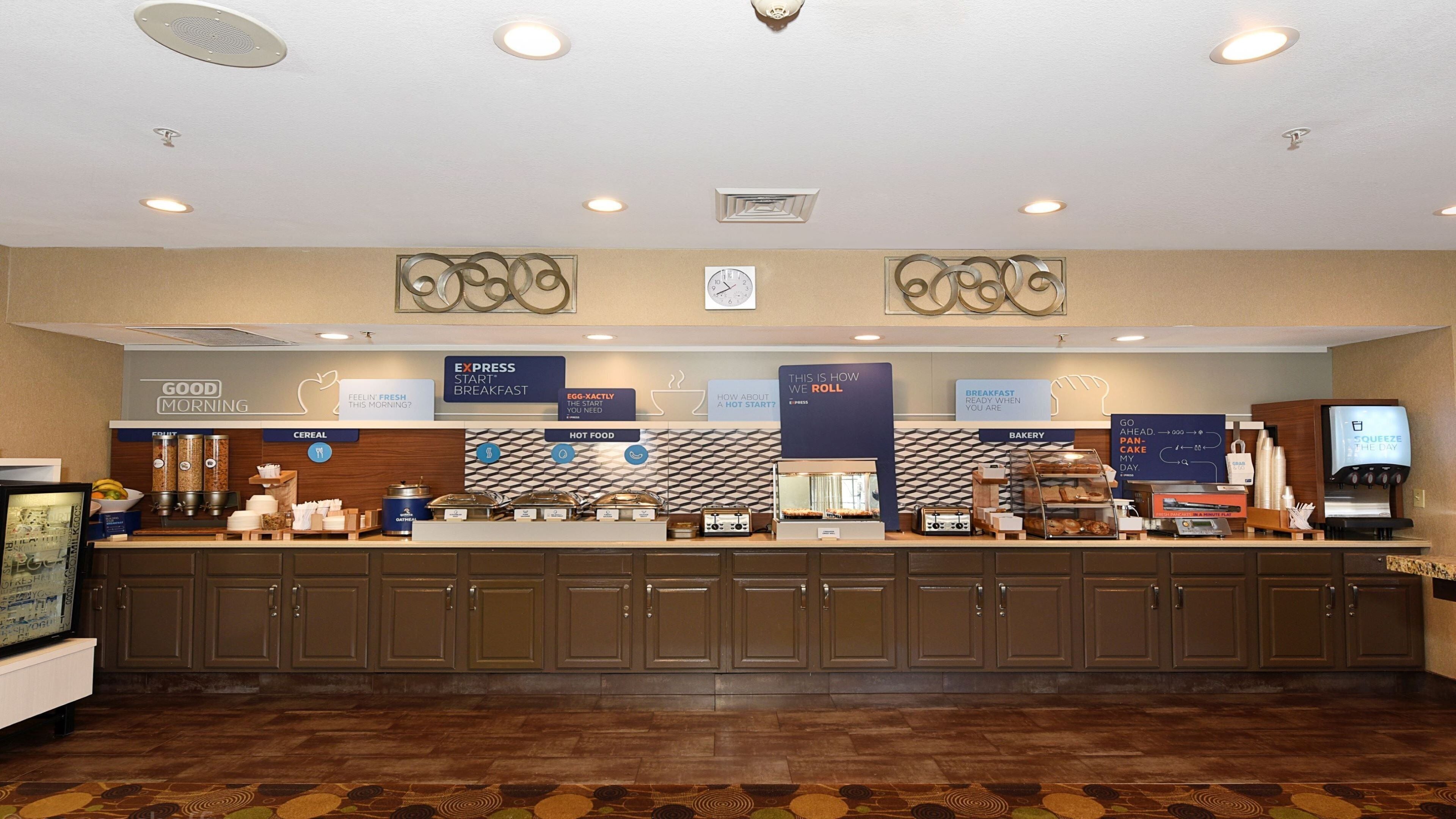 A hotel breakfast area with a counter, breakfast options, and a clock on the wall.