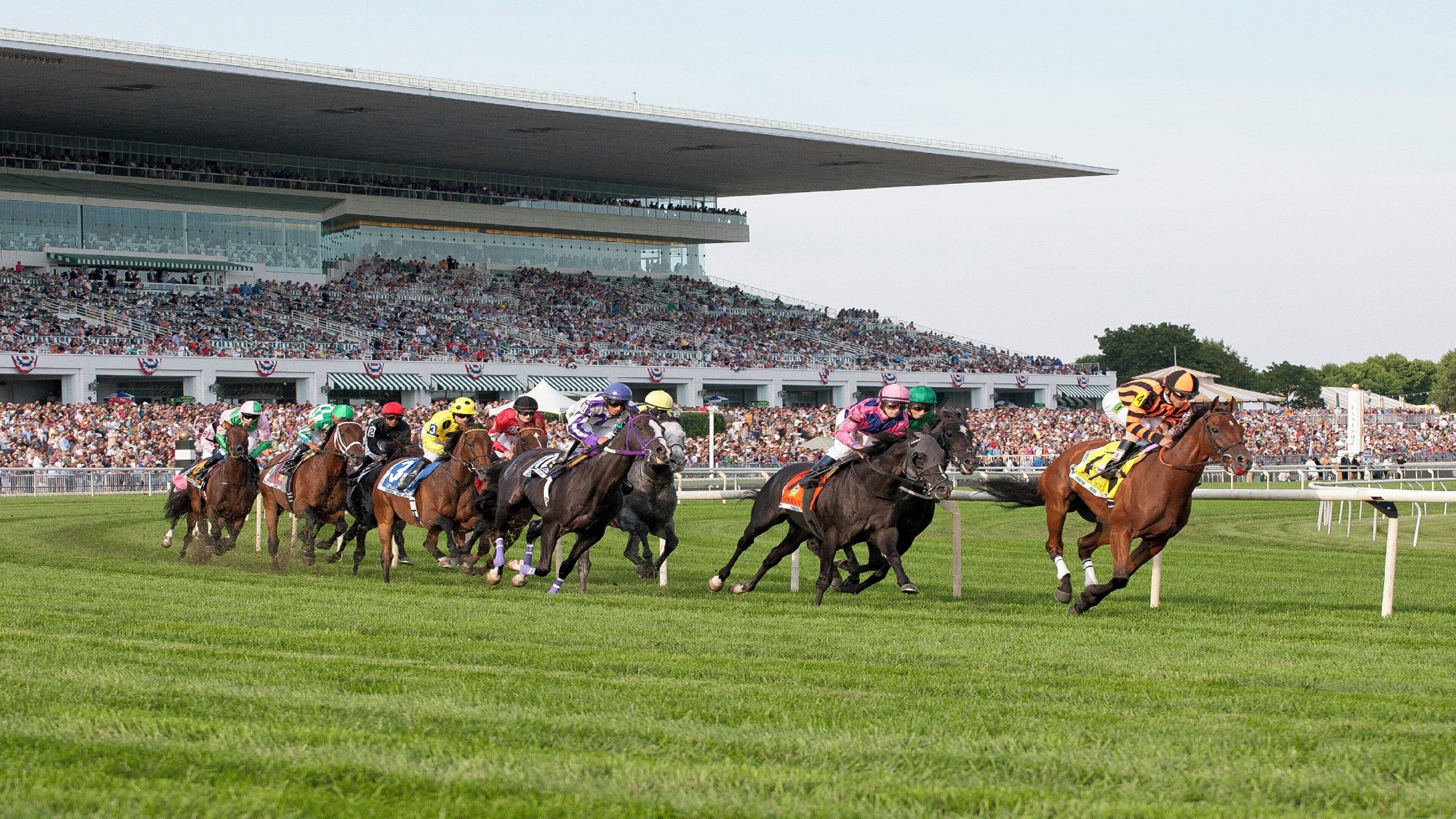 Una carrera de caballos con jinetes, una multitud de fondo y una gran estructura de estadio.