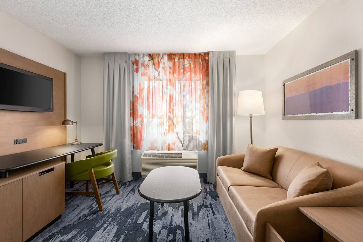 Ein Hotelzimmer mit Flachbildfernseher, Schreibtisch mit Lampe, grünem Sessel, beiger Couch und einem fenster mit Vorhängen.