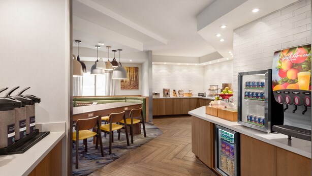 A modern hotel kitchen with a coffee station, a refrigerator with beverages, and a seating area with chairs and a table.