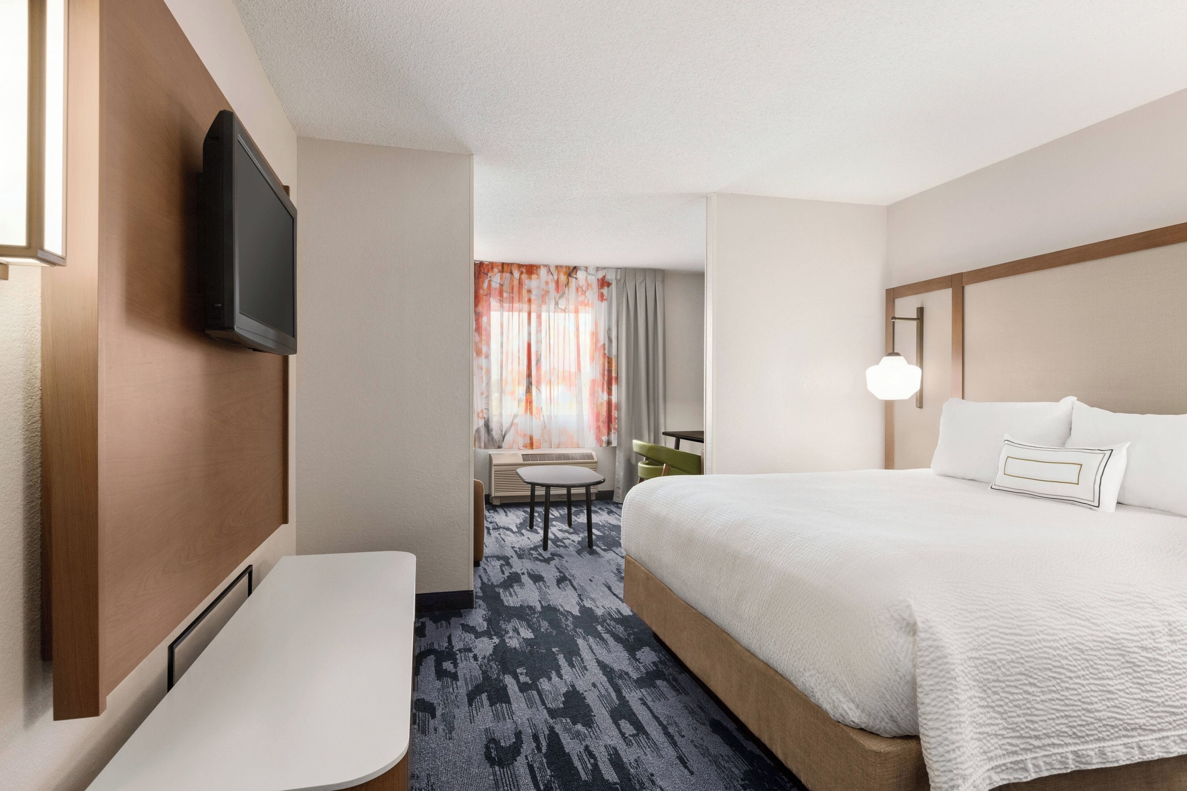 A hotel room with a bed, a desk, a chair, a TV mounted on the wall, and a window with curtains.