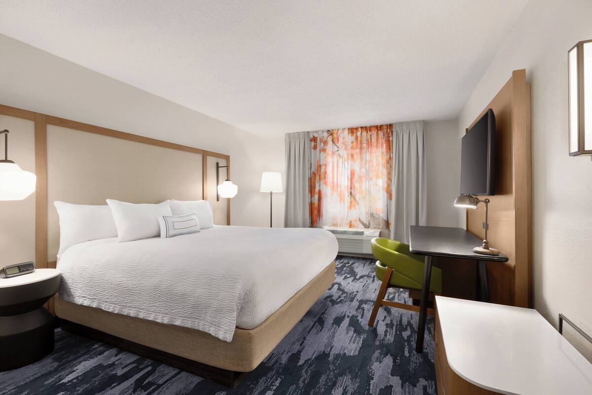 Ein Hotelzimmer mit einem großen Bett, einem Schreibtisch, einem Sessel, einem Fernseher und einem Fenster mit Vorhängen.