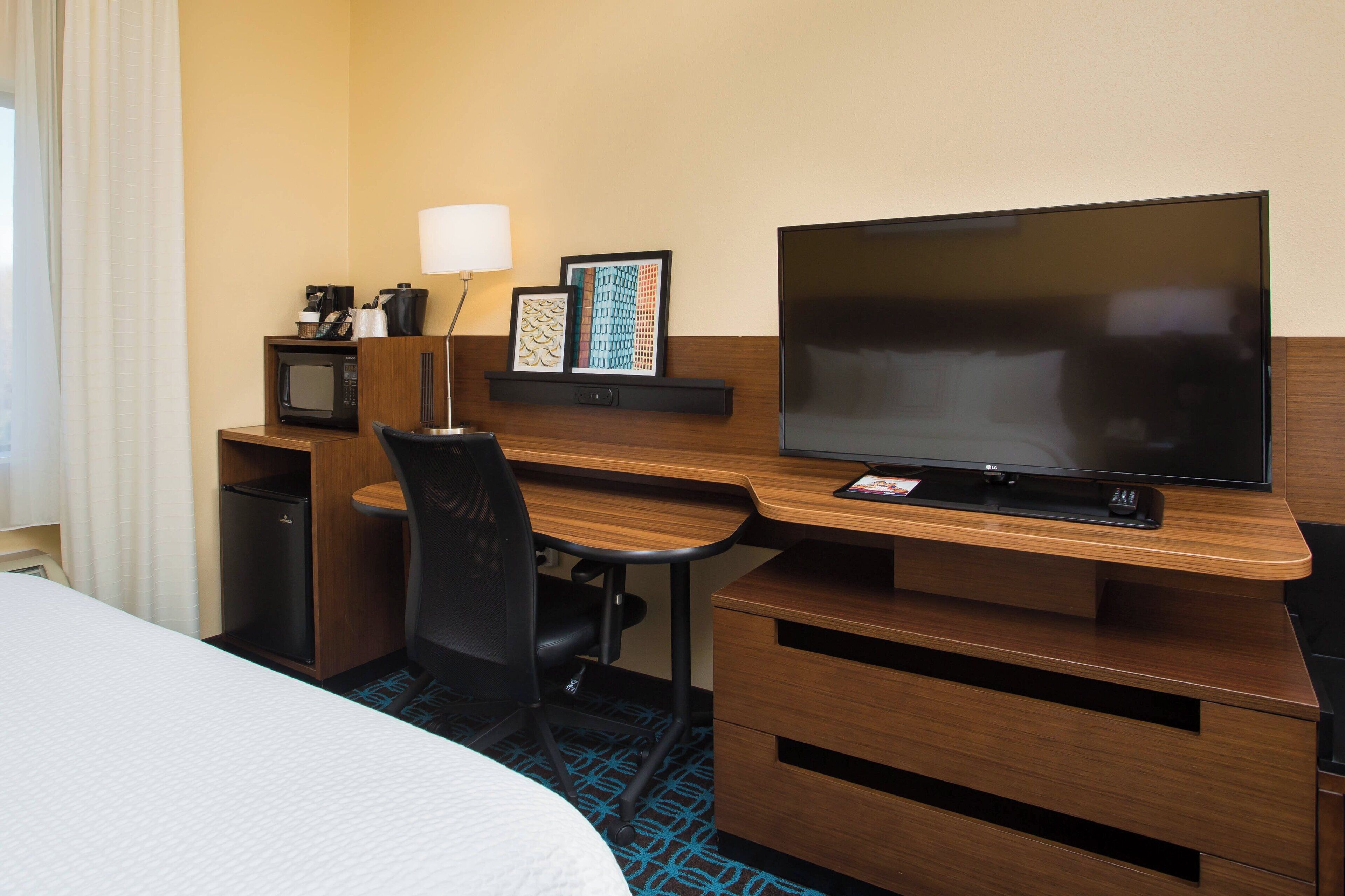 A hotel room with a bed, a desk with a television, a chair, a microwave, a coffee maker, and a small refrigerator.