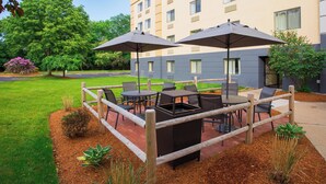 An outdoor patio area with tables and chairs, surrounded by a wooden fence, and a building in the background. - Fairfield by Marriott Inn & Suites Raynham Middleborough/Plymouth (Middleborough)