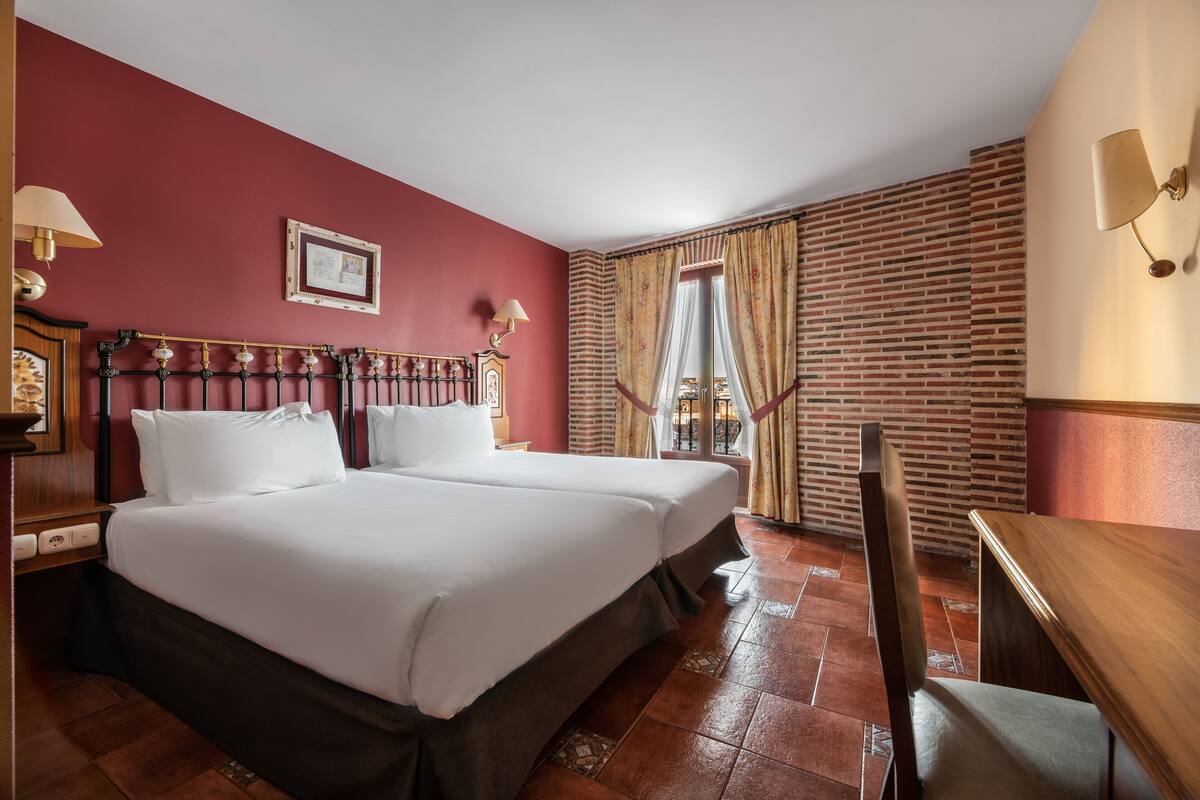 A hotel room with two beds, a wooden desk, a chair, a brick wall, and a window with curtains.