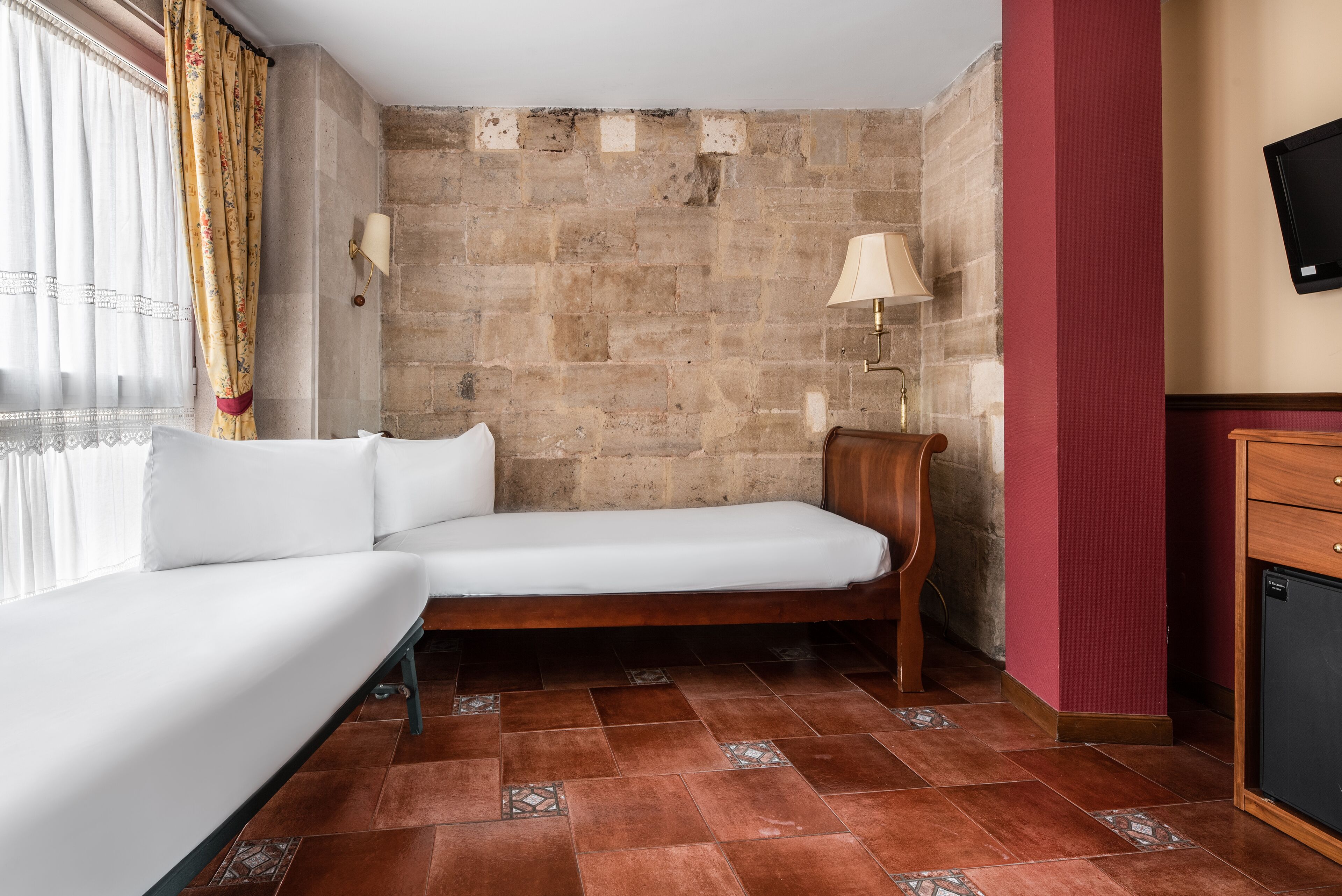 A small, cozy room with a single bed, a wooden nightstand, a wall-mounted TV, and a stone wall.