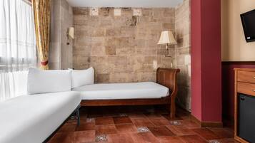 Um quarto pequeno e acolhedor com uma cama de solteiro, uma mesinha de cabeceira de madeira, uma televisão fixada na parede e uma parede de pedra.