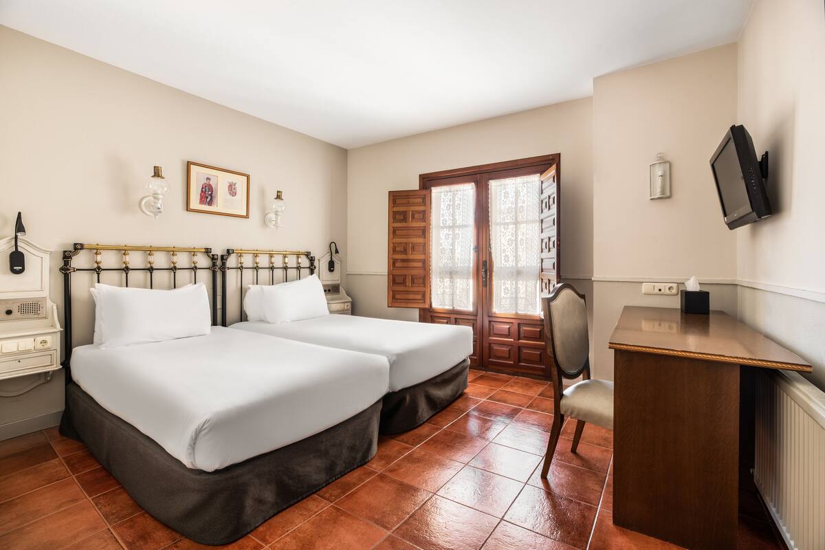 A hotel room with two beds, a desk, a chair, a television, and a window with a view of the outside.
