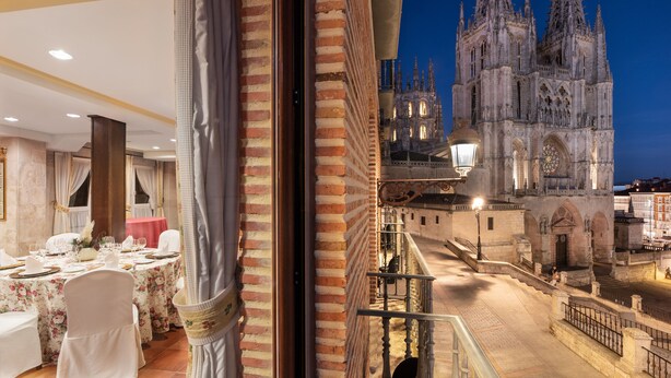 Un comedor con mesas dispuestas para una comida, vista a una catedral con dos torres gemelas y un balcón con barandilla.