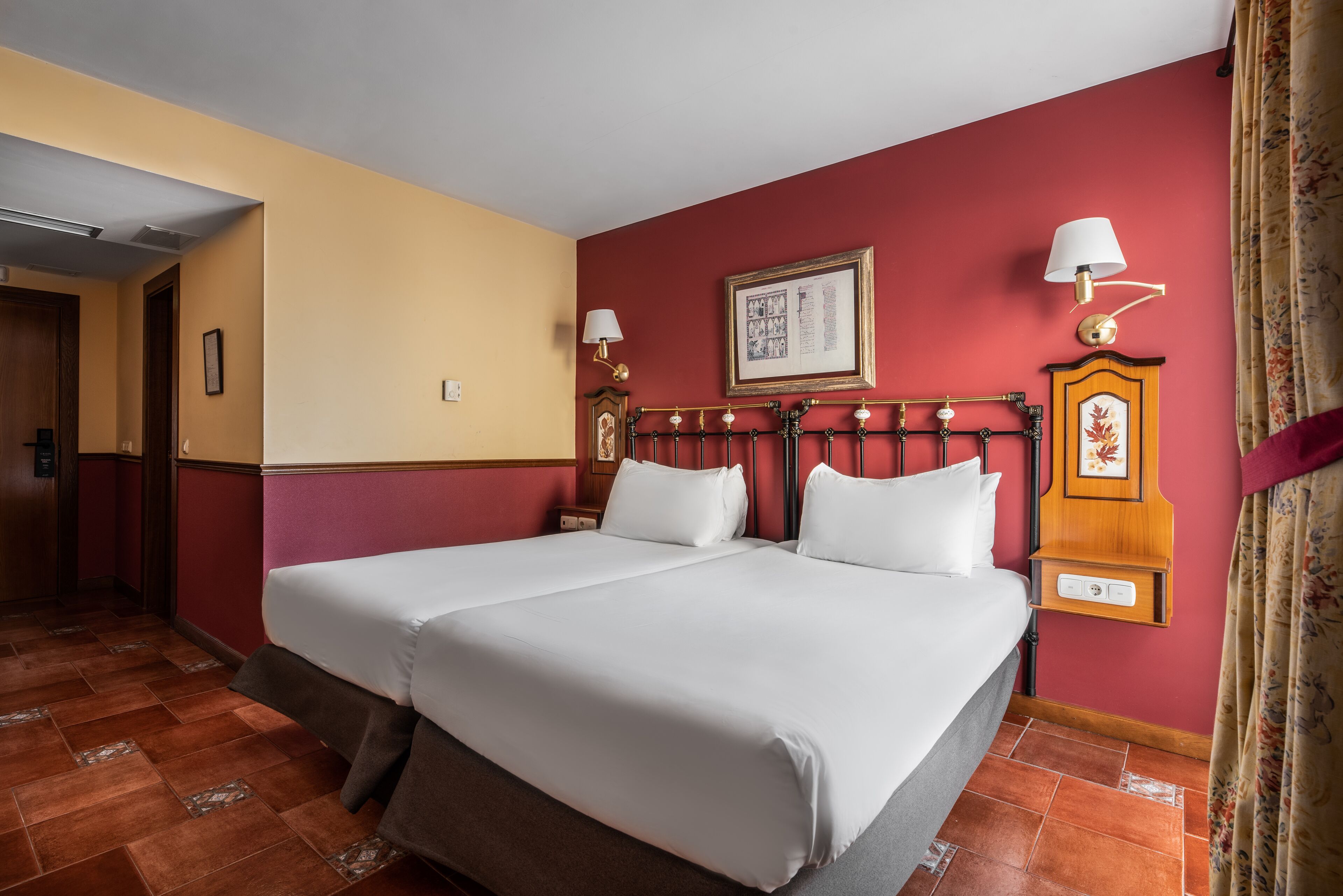 A double bed with white linens, a wooden headboard, two bedside tables with lamps, a framed picture on the wall, and a red curtain.