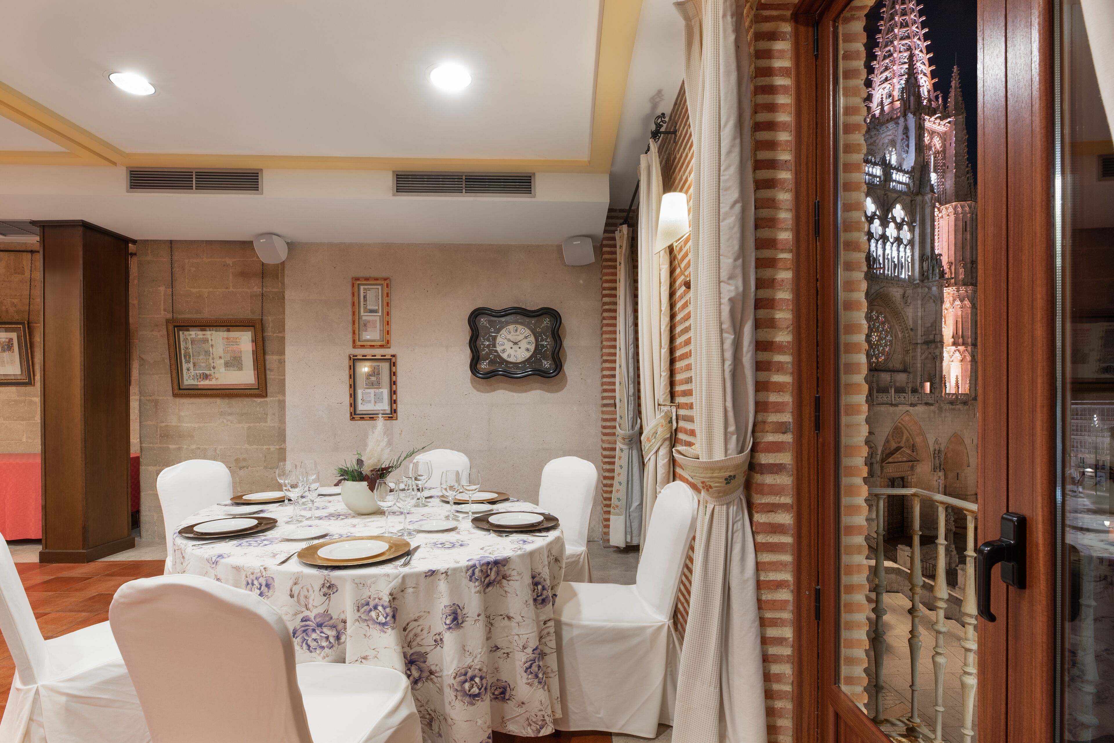 Un coin repas avec une table ronde dressée pour six personnes, des chaises blanches et une nappe à motifs floraux.