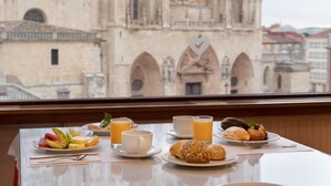 Una mesa de desayuno con platos de fruta, bollería y jugo, y vista a una catedral histórica a través de una ventana.