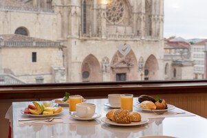 A breakfast table with plates of fruit, pastries, and juice, and a view of a historic cathedral through a window.