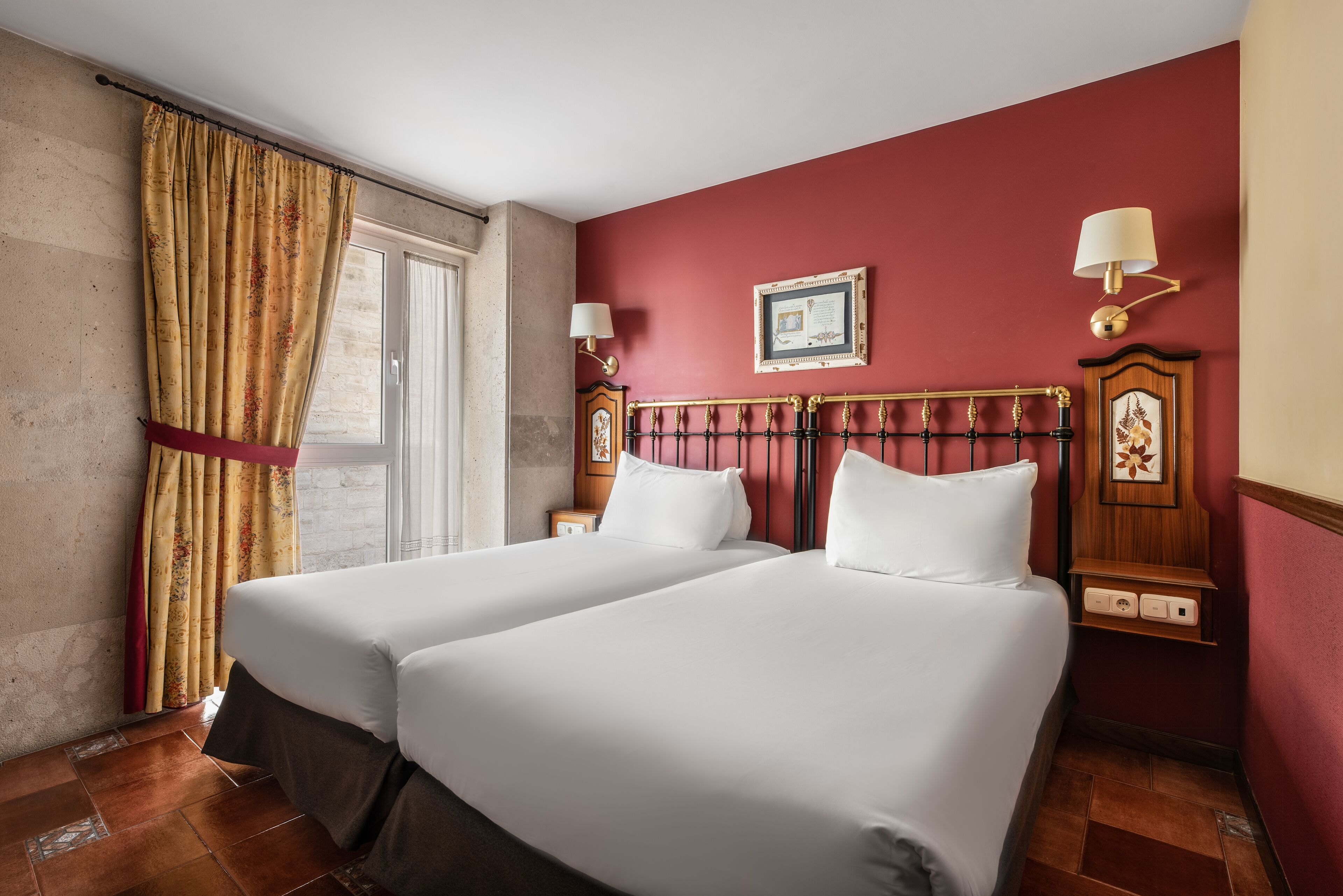 A hotel room with two beds, a window with curtains, a wall-mounted lamp, and a framed picture.