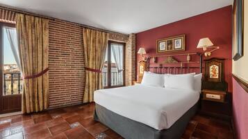 A hotel room with a large bed, a brick wall, a balcony, and a red wall with framed pictures and lamps.