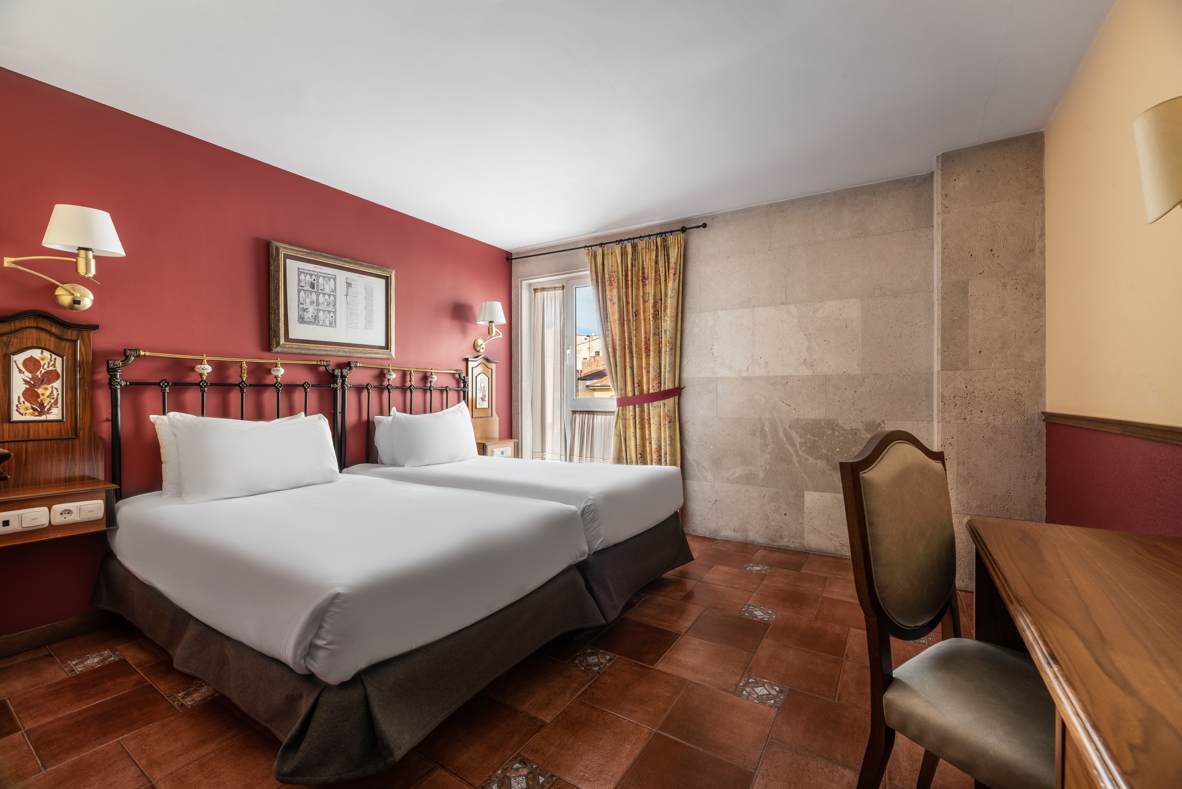 Quarto de hotel com duas camas, uma escrivaninha de madeira, uma cadeira, mesa de cabeceira com luminária e janela com cortinas.