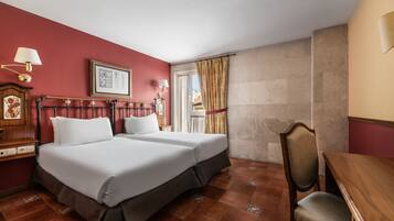 Quarto de hotel com duas camas, uma escrivaninha de madeira, uma cadeira, mesa de cabeceira com luminária e janela com cortinas.