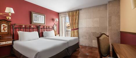Habitación de hotel con dos camas, un escritorio de madera, una silla, una mesita de noche con lámpara y una ventana con cortinas.