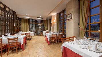 Un comedor con mesas dispuestas para una comida, sillas de madera y ventanas amplias con cortinas.