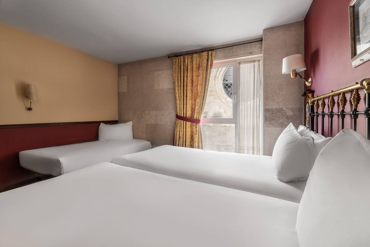 A hotel room with two beds, a window with curtains, a wall-mounted lamp, and a view of the outside.