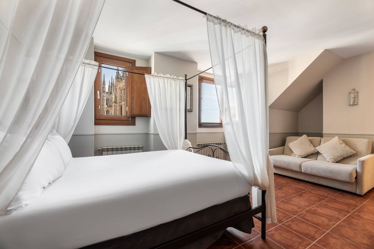 A bedroom with a four-poster bed, a view of a cathedral through a window, a sofa, and a tiled floor.