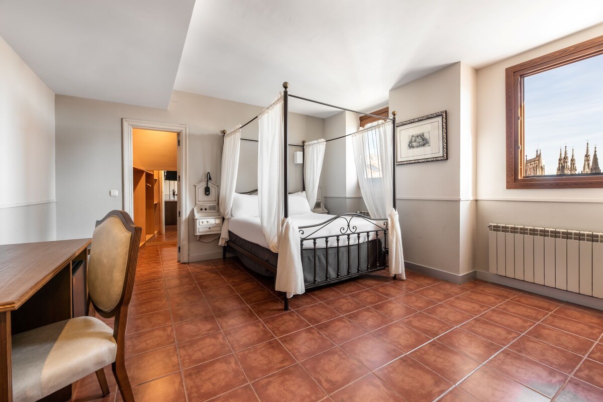 A bedroom with a four-poster bed, a desk with a chair, a radiator, and a window with a view of buildings.