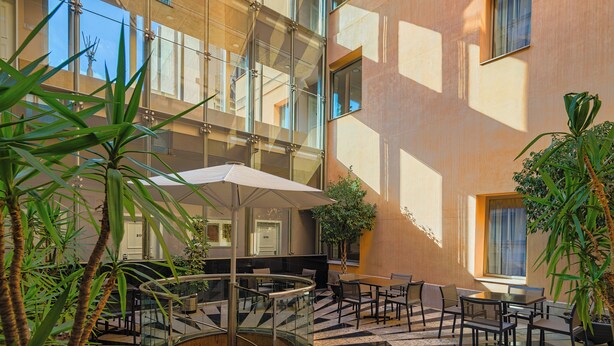 A modern building with a glass atrium, outdoor seating area, and potted plants.
