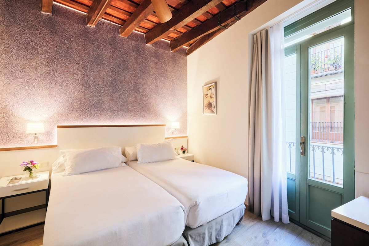 Una cama doble con sábanas blancas, techo de madera, puerta verde y una mesita de noche con lámpara.