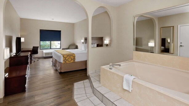 A hotel room with a large bed, a separate desk, a desk chair, a large mirror, a bathtub, and a window with a view of greenery. - Baymont by Wyndham Houston Brookhollow (Houston)