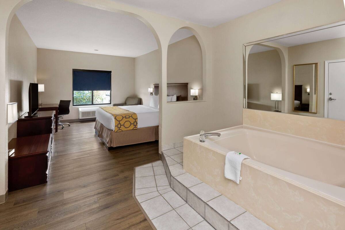 Habitación de hotel con una cama grande, un escritorio independiente, una silla de escritorio, un espejo amplio, una bañera y una ventana con vistas a zonas verdes.