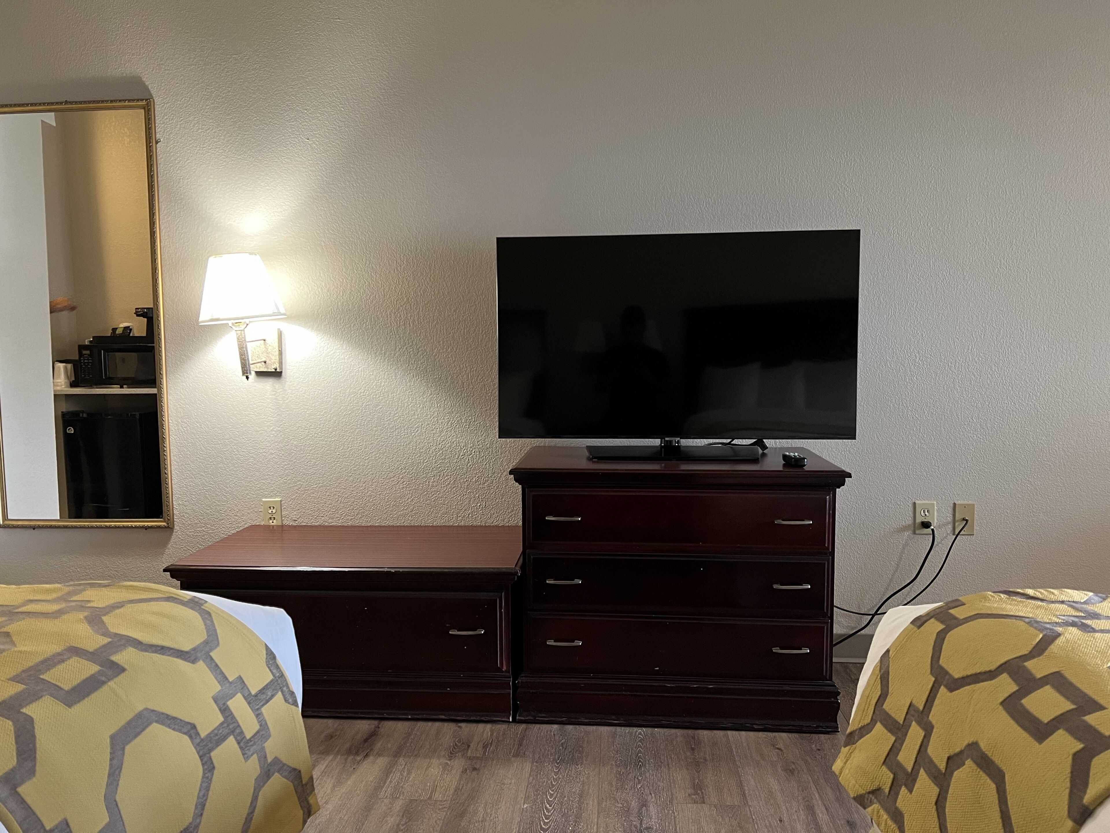 Et hotelværelse med et fladskærms-TV på en trækommode, to senge med mønstret sengetøj, et spejl og en lampe monteret på væggen.