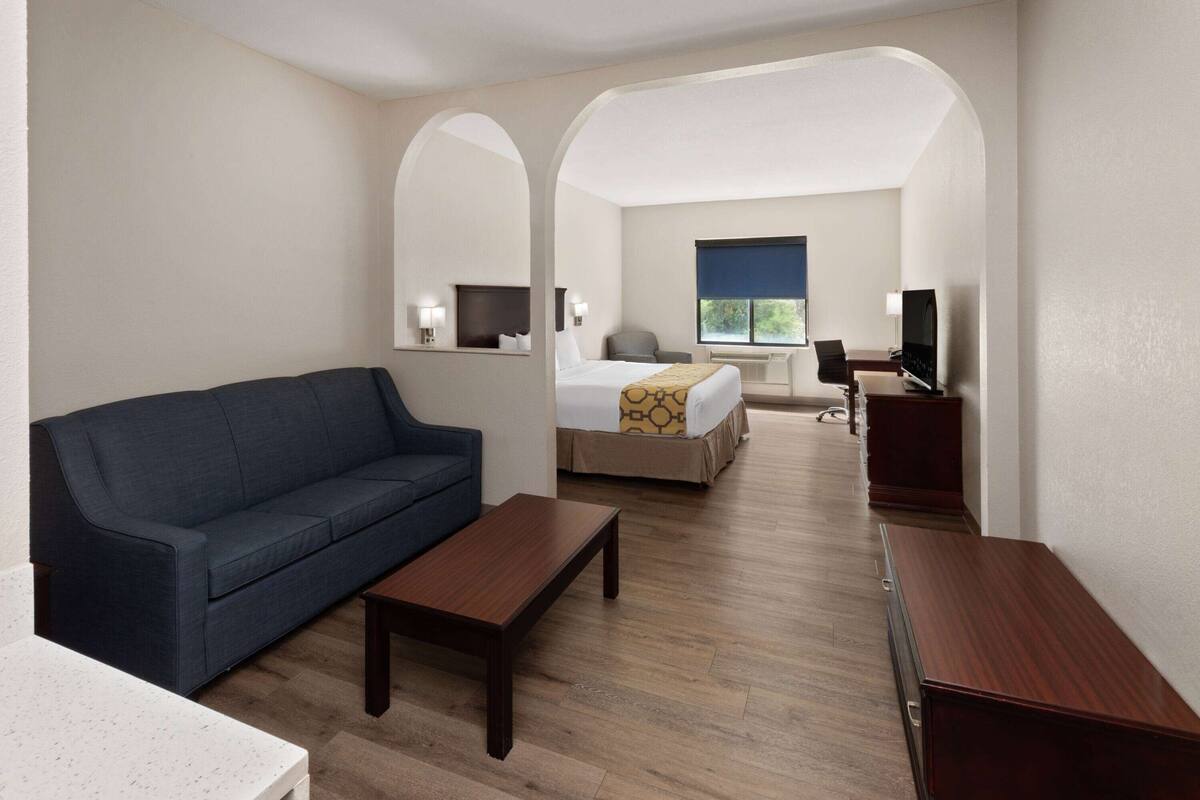 Una habitación de hotel con un sofá azul oscuro, una mesa de centro de madera, una cama con una manta estampada, un escritorio con una computadora y un televisor.