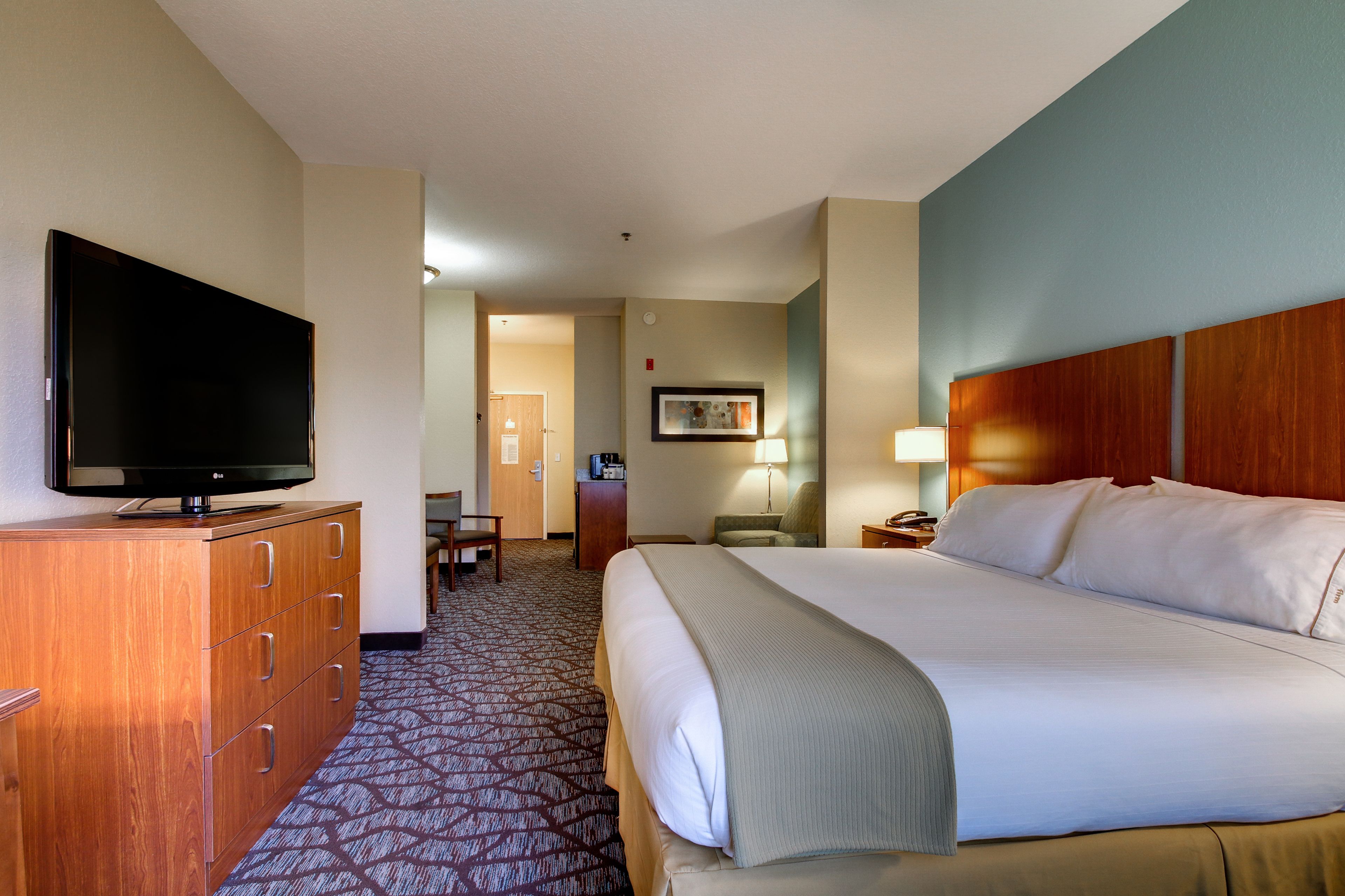 A hotel room with a large bed, a television on a wooden dresser, a desk with a lamp, and a carpeted floor.