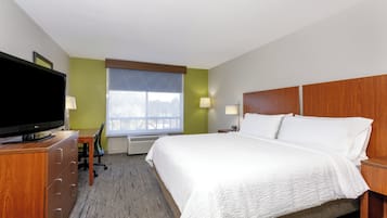 A hotel room with a bed, a desk with a flat-screen TV, a chair, a window with blinds, and a nightstand with a lamp.