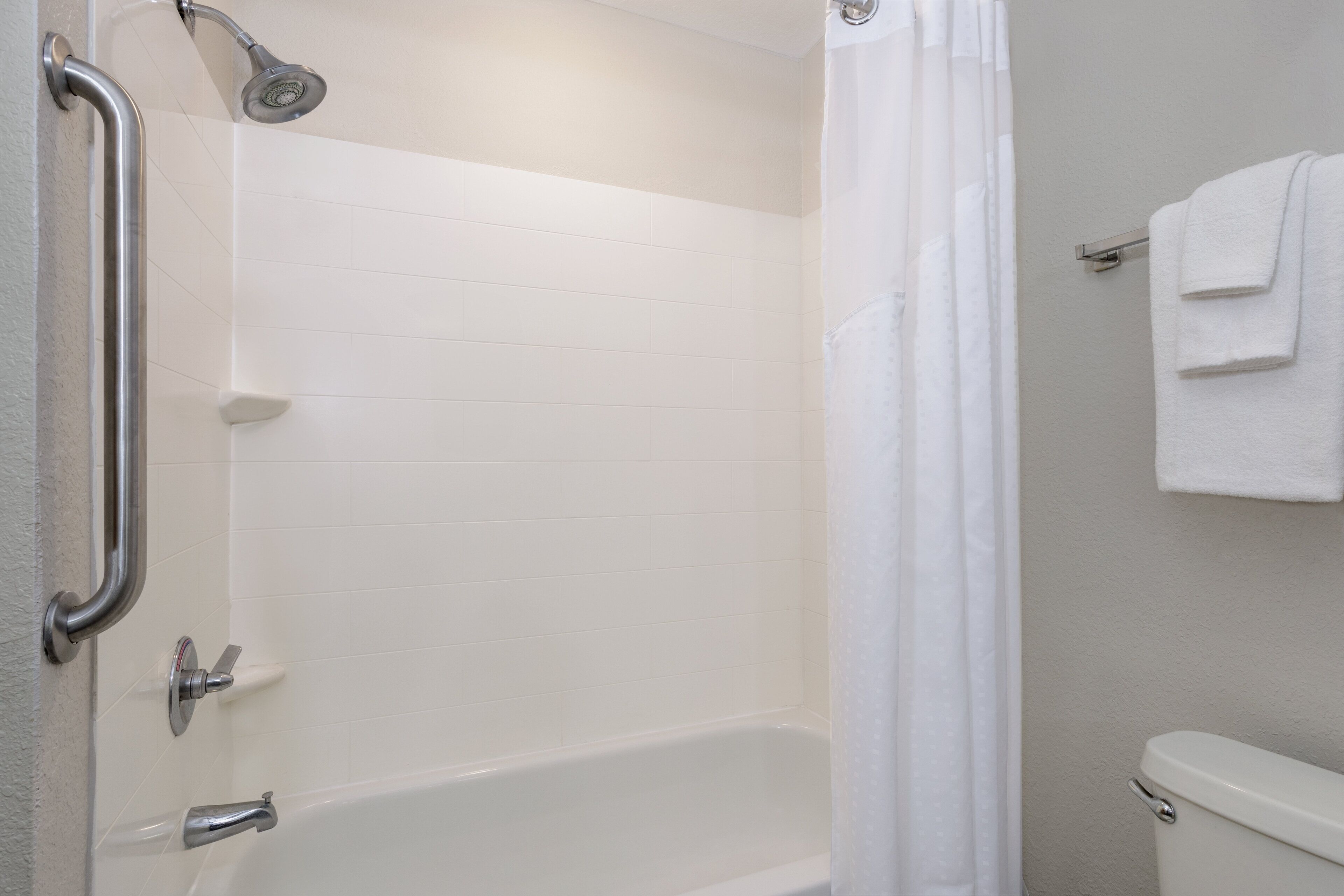A bathroom with a white bathtub, a shower, and a toilet. There is a handrail on the left side and a towel rack on the right.