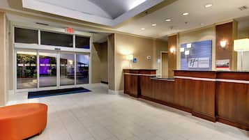 A hotel reception area with a glass door entrance, a reception desk, and a seating area with an orange chair.