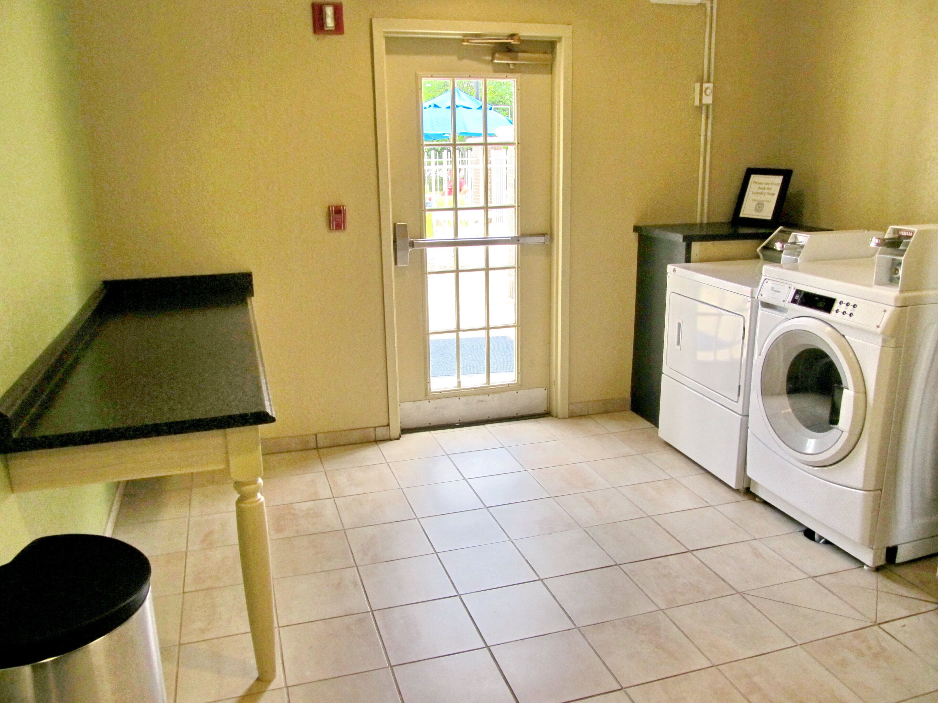 A laundry room with a washing machine and dryer, a door with a view of the outside, and a small trash bin.