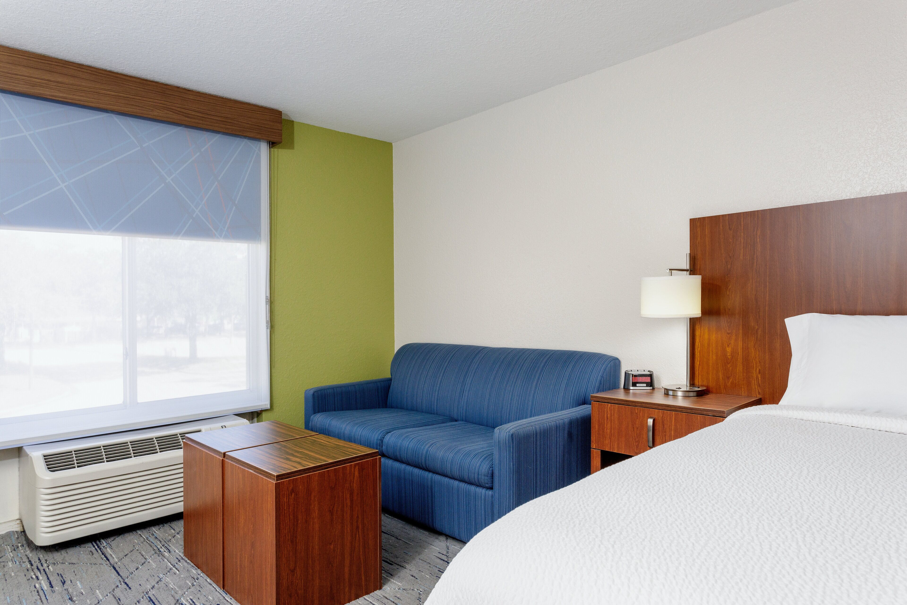 A hotel room with a bed, a seating area with a chair, a bedside table with a lamp, and a window with blinds.