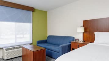 A hotel room with a bed, a seating area with a chair, a bedside table with a lamp, and a window with blinds.